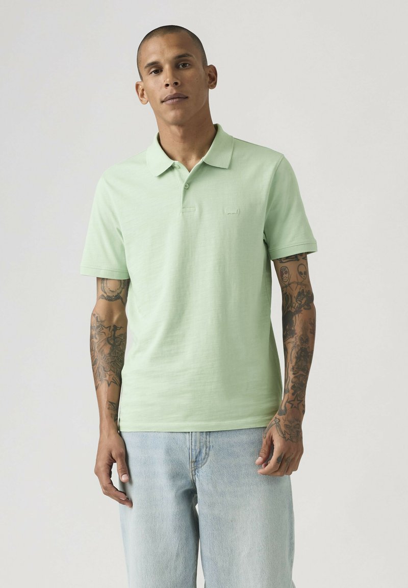 Man met getatoeëerde armen draagt een lichtgroen poloshirt en lichtblauwe jeans, staand tegen een effen achtergrond.