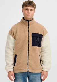 Chaqueta de forro polar en color beige y crema con un bolsillo negro. Presenta un cuello alto, cierre con cremallera y una textura suave. Detalle de diseño utilitario.