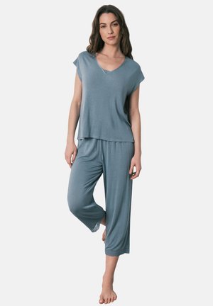 Kvinde står barfodet i løstsiddende, lyseblåt pysjamasæt med kortærmet V-udskæring og korte bukser med vide ben mod en ensfarvet baggrund.