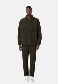 Giacca in felpa verde scuro con collo a camicia, due tasche frontali e chiusura con bottoni, abbinata a pantaloni coordinati e sneakers bianche.