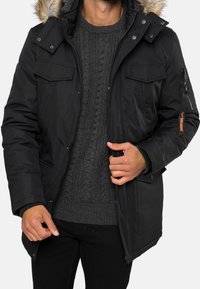 Sort parka-jakke med en pelsforet hætte, der har lynlåse og knap-lukkede lommer, struktureret strikket sweater underneath, i en afslappet pasform.