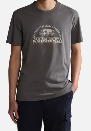 Mężczyzna w szarej koszulce z krótkim rękawem z logo "Antarctic Research Program Napapijri Geographic" i ciemnych spodniach typu cargo, z jedną ręką w kieszeni.