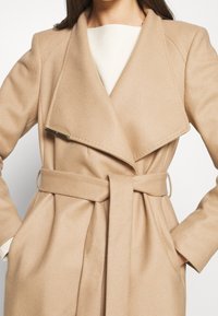 Vrouw draagt een beige wollen mantel met een ceintuur en brede revers over een witte trui, handen in de mantelzakken gestopt.