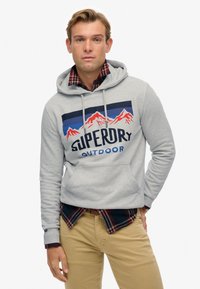 Felpa grigia con logo frontale, grafica di montagne in rosso e blu, tasca kangaroo, indossata sopra una camicia a quadri e pantaloni beige.