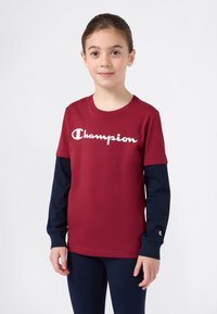 Jente iført en rød Champion T-skjorte over en langermet marineblå skjorte og marineblå bukser, stående mot en ensfarget lys bakgrunn.