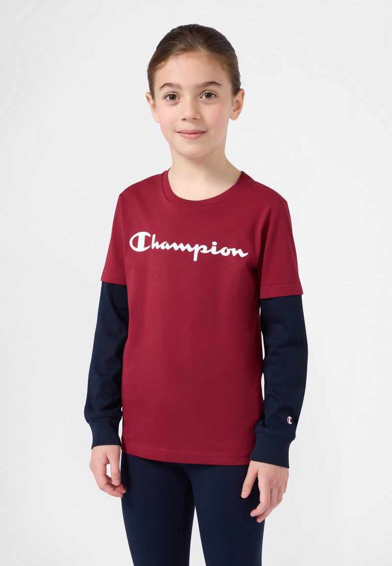 Jente iført en rød Champion T-skjorte over en langermet marineblå skjorte og marineblå bukser, stående mot en ensfarget lys bakgrunn.