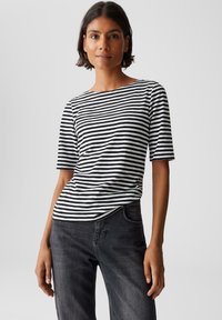 Femme portant un t-shirt rayé horizontal noir et blanc à manches courtes et un jean gris foncé, se tenant devant un fond clair uni.
