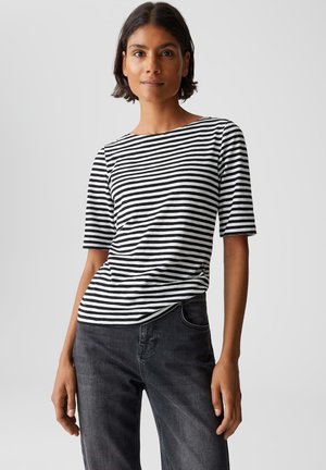 Vrouw in een zwart/wit horizontaal gestreept shirt met korte mouwen en donkergrijze jeans, staand tegen een effen lichte achtergrond.
