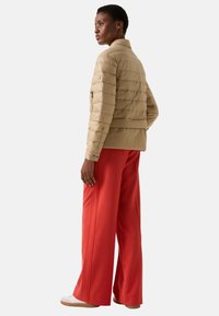 Beżowa pikowana kurtka puchowa z poziomym pikowaniem, mankietami na rękawach i bocznymi kieszeniami na zamek, zestawiona z jasnoczerwonymi szerokimi spodniami.