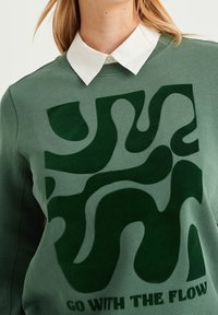 Zelená bavlněná mikina s výrazným abstraktním tmavě zeleným vzorem a textem "JDI S TOKEM" v černé barvě, pod kterou je bílý límec.