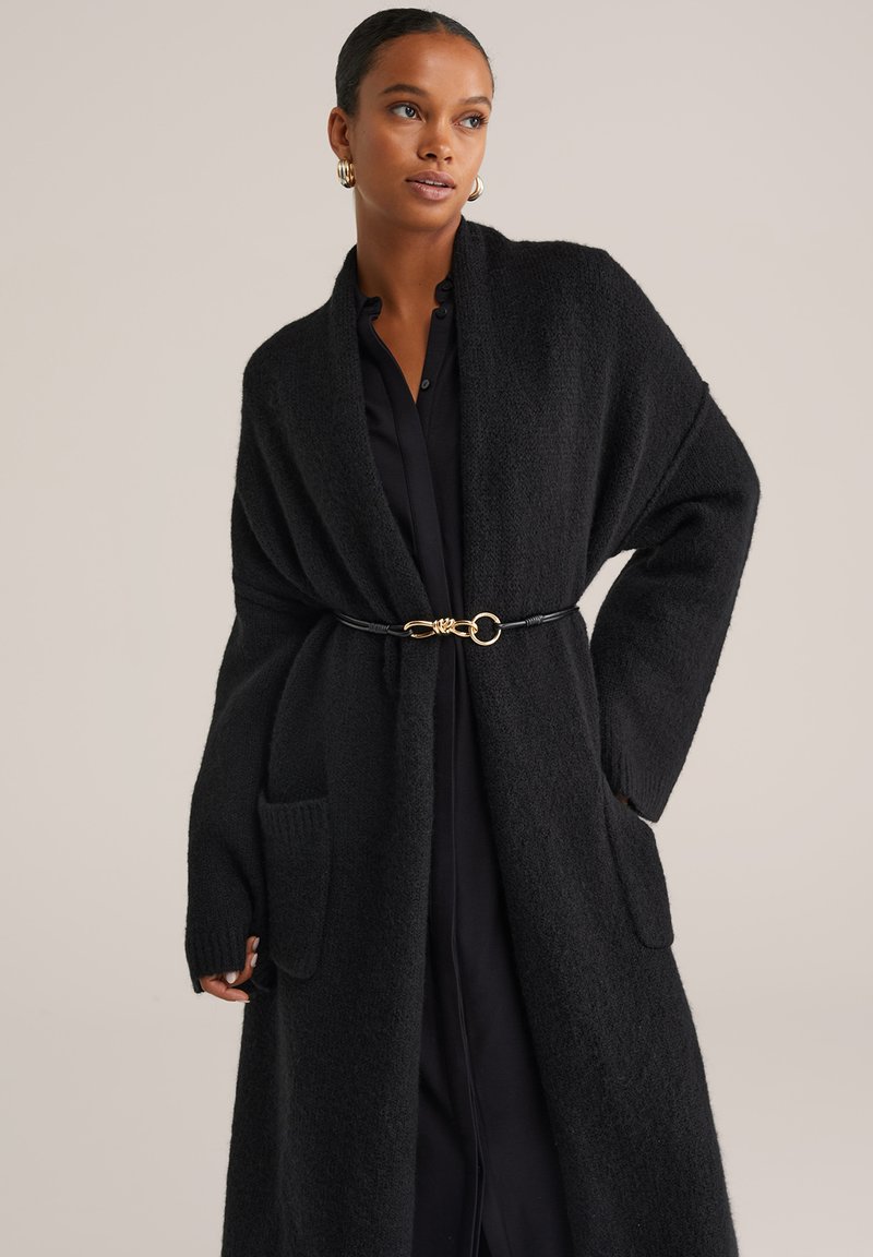 Žena oblečená v dlhom čiernom textúrovanom kardigane s prednými vreckami, opásaná v páse, cez čierne šaty na gombíky, pózujúca na jednoduchom pozadí.