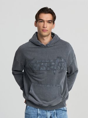 Felpa grigia in cotone con grande logo "waste" in rilievo, tasca frontale, vestibilità comoda e tessuto dalla trama strutturata. Vista frontale, modello indossa jeans strappati.