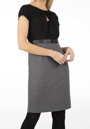 Vrouw draagt een zwart kortemouwjurk met een grijs rokgedeelte, een zwarte tailleband, zwarte panty's en een zilveren armband om haar rechterpols.