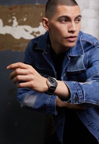 Montre noire avec un cadran rond et des accents argentés sur un bracelet noir, portée sur une main avec une veste en denim de couleur bleu délavé.
