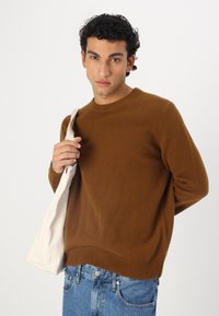 Pull en tricot marron avec un col rond, des poignets et un ourlet côtelés ; associé à un jean en denim bleu et un sac à épaule en tissu léger.