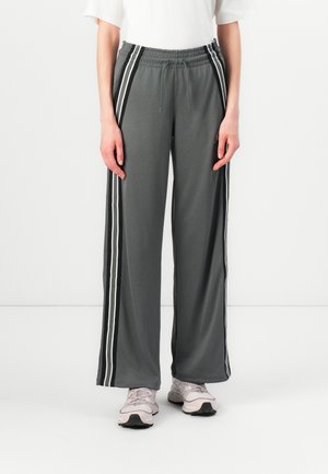 Pantaloni a gamba larga grigi con una morbida texture, caratterizzati da strisce laterali bianche e una cintura elastica. Abbinati a scarpe da ginnastica chiare.