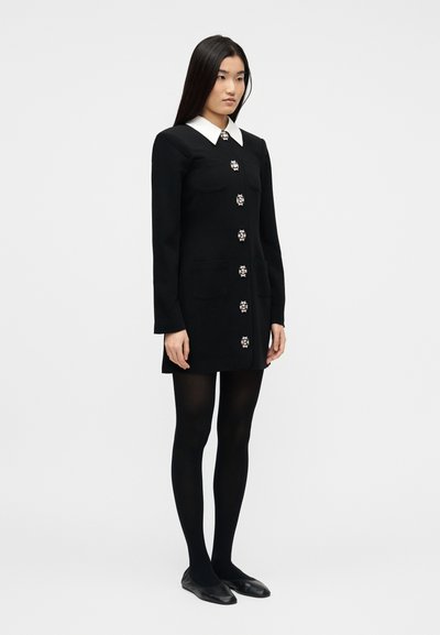 Γυναίκα με μαύρο φόρεμα μακρυμάνικο και λευκό γιακά, διακοσμητικά κουμπιά, μαύρες καλσόν και μαύρες μπαλαρίνες, στέκεται μπροστά σε απλό φόντο.