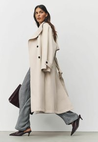 Beige trench coat with black buttons, wide lapels, and a tied belt. Paired with gray textured trousers and burgundy mules, featuring a structured handbag.