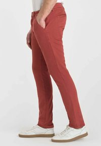 Man som står med ena handen i fickan, klädd i slim fit röda byxor och vita sneakers med gummisula mot en enfärgad bakgrund.