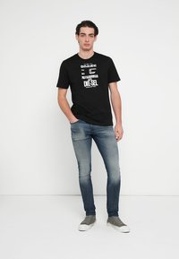 Camiseta negra de algodón con texto impreso en blanco y negrita, combinada con vaqueros ajustados de color azul claro y zapatillas grises con suelas blancas.