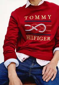 Red sweatshirt with embroidered "TOMMY HILFIGER" logo and rope design. Worn over a light blue collared shirt. Dark blue jeans visible.