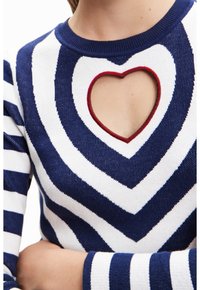 Granatowy i biały sweter w paski z sercowym wycięciem na wysokości klatki piersiowej, obramowanym na czerwono, noszony przez osobę z założonymi na sobie rękami.
