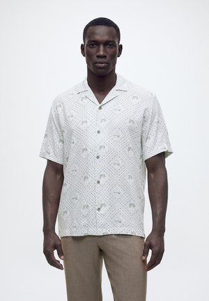 Άνδρας που φοράει λευκό πουκάμισο με κοντά μανίκια και κουμπιά, με σχέδια φοίνικων και διαμαντιών, συνδυασμένο με μπεζ παντελόνι, στέκεται μπροστά σε απλό φόντο.