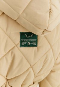 Béžová prošívaná bunda s zelenou visačkou s bílým textem a minimalistickou grafikou. Tkanina působí měkce a má jemný lesk.