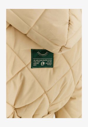 Beżowa pikowana kurtka z zieloną metką z białym napisem i minimalistyczną grafiką. Tkanina wydaje się miękka z lekkim połyskiem.