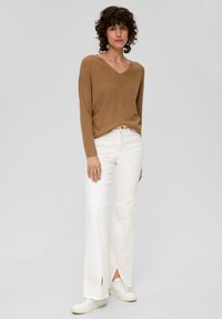 Femme avec des cheveux bouclés portant un pull brun à col en V, un pantalon blanc à ourlet fendu et des baskets blanches, se tenant contre un fond uni.