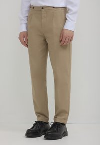 Beige Hosen mit glatter Textur, versehen mit zwei vorderen Falten und einem standardmäßigen Bund mit schwarzen Knöpfen. Kombiniert mit schwarzen Schnürschuhen.