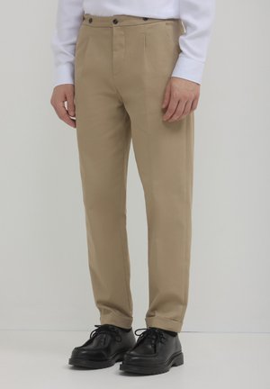 Beige Hosen mit glatter Textur, versehen mit zwei vorderen Falten und einem standardmäßigen Bund mit schwarzen Knöpfen. Kombiniert mit schwarzen Schnürschuhen.
