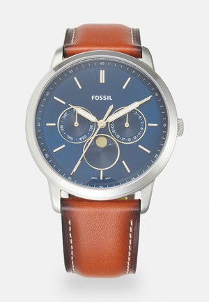 Montre avec un bracelet en cuir marron, un cadran bleu, un boîtier en acier inoxydable argenté, des accents dorés et plusieurs sous-cadrans pour le jour et la date.