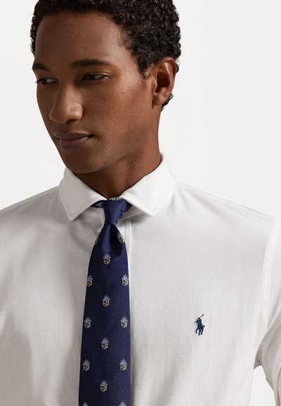 Λευκό πουκάμισο με κολάρο, διακοσμημένο με ένα μικρό μπλε λογότυπο polo. Συνδυασμένο με μια ναυτική γραβάτα, με σχέδιο που περιλαμβάνει χρυσές και μπλε κρητικές παραστάσεις.