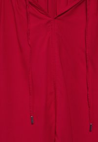 Tuta rossa con scollatura a V, dotata di corde di regolazione e accenti metallici. Il tessuto appare morbido e leggero con un colore uniforme.
