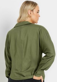 Chemise légère de couleur vert olive, à manches longues, avec un col structuré, des poignets boutonnés et un détail de couture au dos. Texture lisse, coupe décontractée.