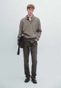 Brązowy sweter dzianinowy z kołnierzem i guzikami, noszony na koszuli w kratę; ciemnozielone dżinsy i czarne buty; trzyma czarną torbę.