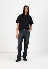 Black short-sleeve t-shirt, relaxed-fit dark blue jeans, and black sneakers with white accents, posing against a plain white background.