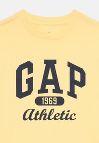 Gul t-skjorte i bomull med svart fet "GAP" tekst, "1969" i mindre skrift, og "Athletic" i kursiv. Rund hals, korte ermer.