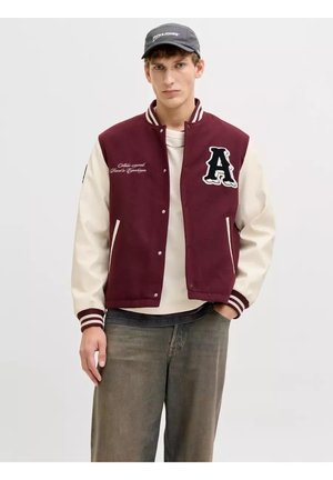 Giovane uomo che indossa una giacca varsity bordeaux e bianca con una toppa a forma di "A", una maglietta bianca, jeans grigi e un cappello nero Jack & Jones.