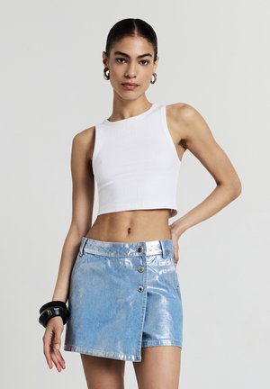Young woman with slicked-back hair wearing a white ribbed crop top, metallic blue mini skirt with buttons, large hoop earrings, and black bracelets.