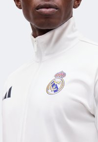 Gros plan d'une personne portant une veste de sport blanche zippée avec le logo du Real Madrid et des bandes Adidas sur la poitrine.