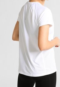 Camiseta deportiva blanca de manga corta, fabricada con tejido de malla transpirable, que presenta un ajuste holgado y una sutil marca en el dobladillo.