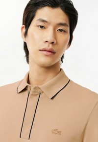 Beżowa koszulka polo z teksturowanej tkaniny, z kontrastowym granatowym wykończeniem kołnierzyka oraz małym haftowanym logo na piersi. Dopasowany krój.