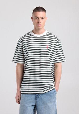 Jeune homme debout, portant un t-shirt blanc et noir à rayures horizontales, manches courtes, avec un petit logo homard rouge sur la poitrine et un jean bleu clair.