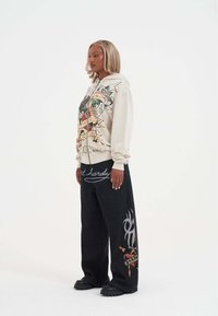 Beżowa bluza z kolorowym nadrukiem, zestawiona z czarnymi spodniami o szerokim kroju z wyszukanymi wzorami oraz dużymi czarnymi butami.