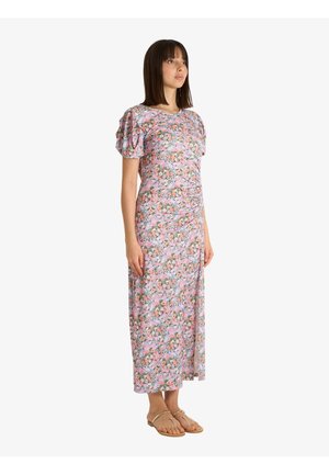 Donna in piedi che indossa un vestito rosa a fiori con maniche a sbuffo e uno spacco laterale, abbinato a sandali piatti beige su sfondo bianco.