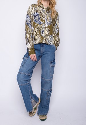 Vrouw draagt een olijfgroen paisley-blouse, gestopt in een blauwe denim cargobroek en beige sneakers, staand tegen een effen witte achtergrond.