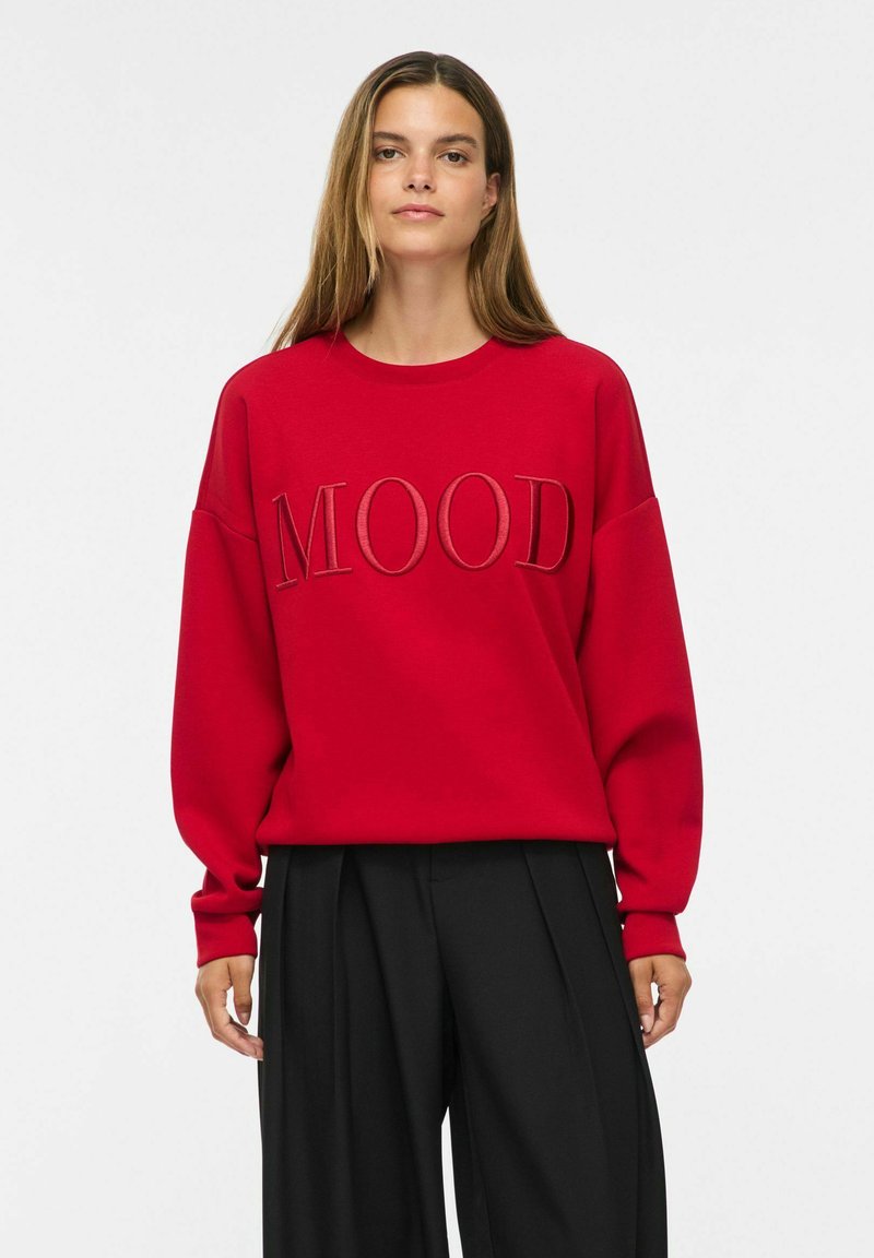 Femme aux longs cheveux portant un sweat rouge avec le mot "MOOD" en grandes lettres et un pantalon noir ample, debout devant un fond uni.