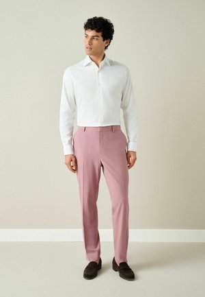 Mann steht drinnen, trägt ein weißes Hemd, pinke Hose und dunkelbraune Loafers und schaut seitlich an eine beige Wand.